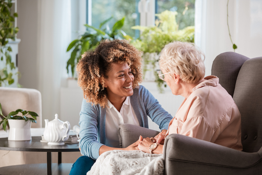 African American female communicating with senior woman living with dementia, sitting in living room holding hands and listening to her carefully