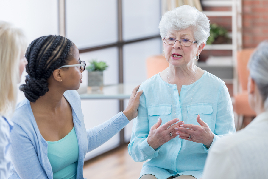 Mid-adult African American female caregiver comforts a senior Caucasian woman. The senior woman is discussing how to navigate her husband’s Alzheimer’s’ disease during a support group at a retirement community